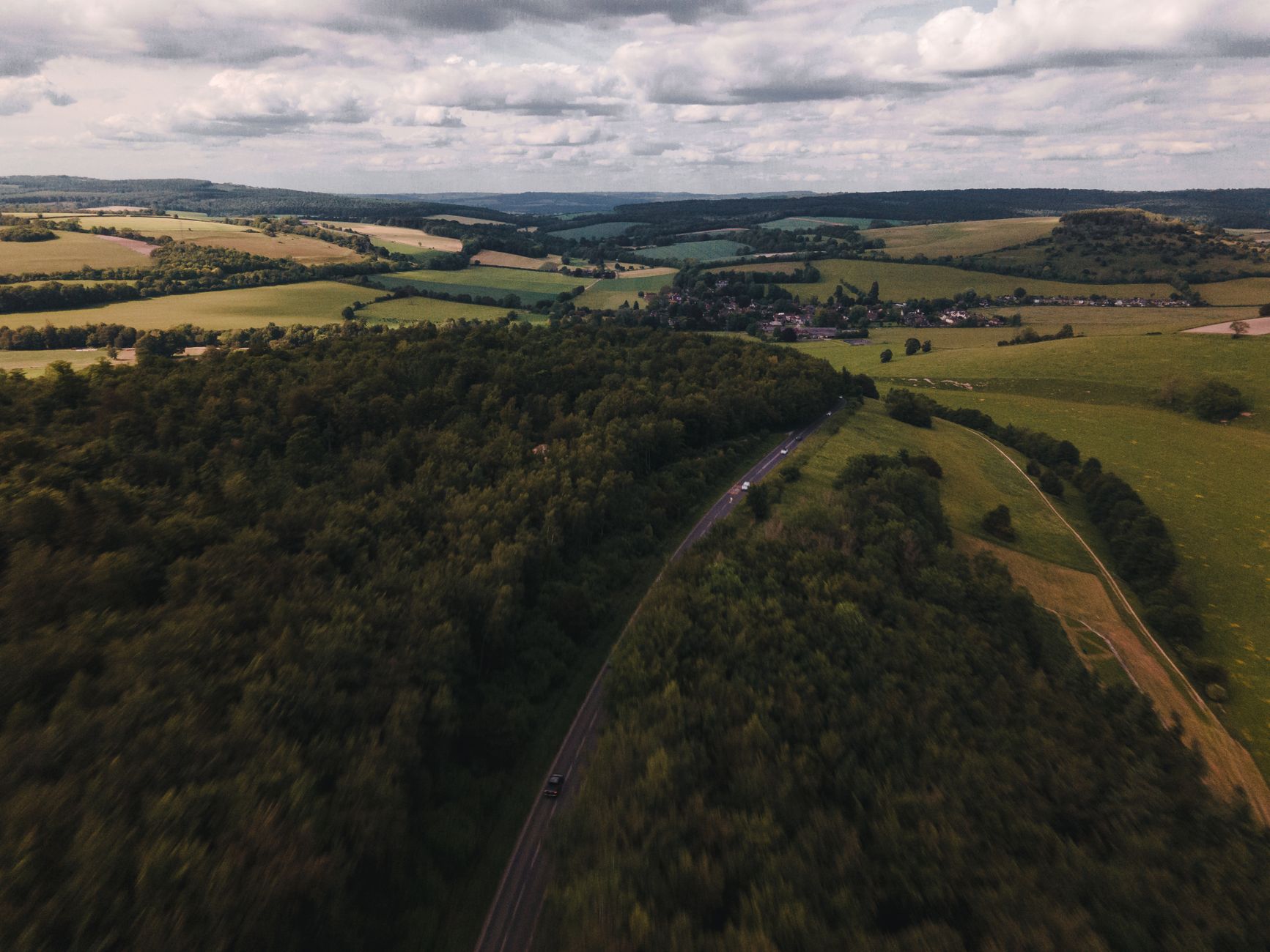 Aerial view of rolling countryside fields, hedgerows, and a winding road leading to a small village.