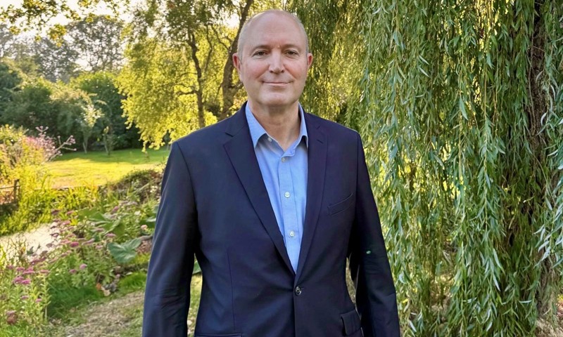 Bald man in a blue shirt and dark suit standing outdoors beside a willow tree.