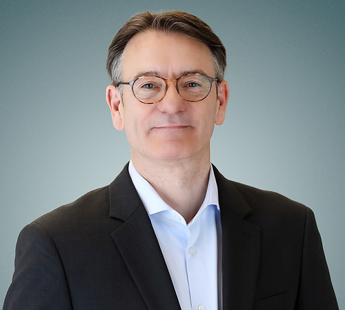 Smiling man in a dark suit and blue shirt wearing round glasses against a light blue background.