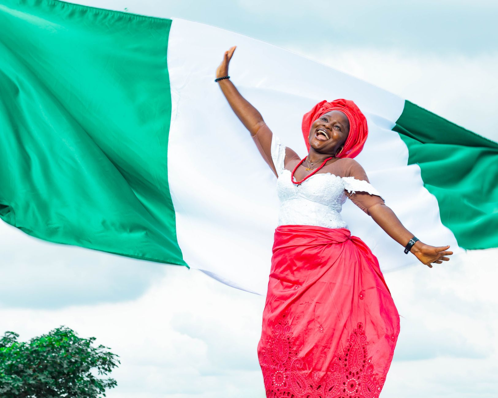 Smiling woman in red headscarf and dress waves arms beneath large green-white flag outdoors.